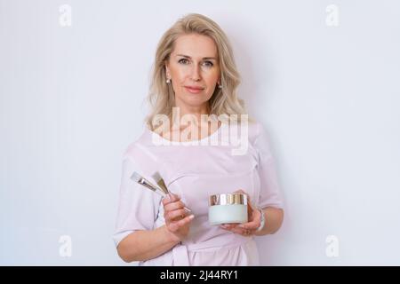 An adult woman beautician dermatologist holds brushes for creams and masks on a light background in the office and smiles. Professional facial skin care in a beauty salon Stock Photo