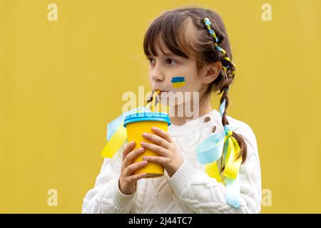 A little Ukrainian girl with two pigtails and yellow and blue ribbons drinks a hot drink from a glass of patriotic colors like the Ukrainian flag. Stop the war in Ukraine. Volunteer Help for Refugees. High quality photo Stock Photo