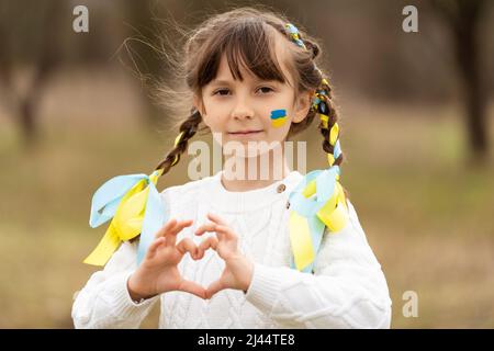 A beautiful little girl with pigtails and braided yellow and blue ribbons, like the color of the Ukrainian flag, makes a heart with her hands. I love Ukraine. Stop the war. . High quality photo Stock Photo