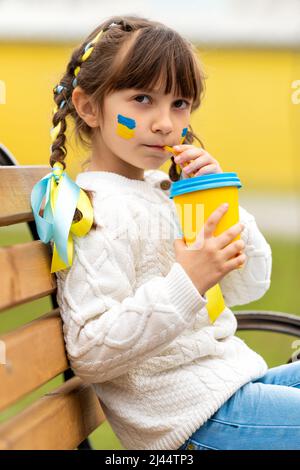 A little Ukrainian girl with two pigtails and yellow and blue ribbons drinks a hot drink from a glass of patriotic colors like the Ukrainian flag. Stop the war in Ukraine. Volunteer Help for Refugees. High quality photo Stock Photo