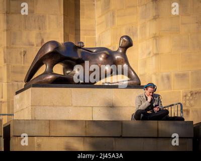 Young male sitting cross-legged in front of a bronze Henry Moore statue, looking down at his phone. Leeds Art Gallery. UK Stock Photo