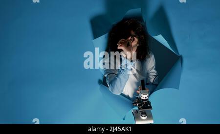 Crazy foolish scientist using laboratory microscope in studio, being silly and doing goofy funny expressions. Mad amusing chemist looking insane after substance explosion, messy hair. Stock Photo