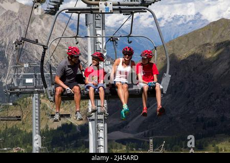 Montgenevre (French Alps, south-eastern France): family sitting on a chairlift going to the mountain biking trails. Parents with two children, wearing Stock Photo