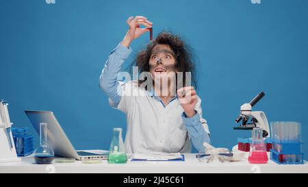 Insane foolish female chemist working with microscope in studio, acting crazy and insane on camera. Goofy amusing woman feeling silly and being funny, mad playful scientist with messy hair. Stock Photo