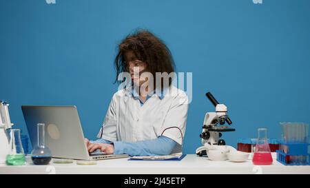 Portrait of crazy female chemist working on laptop at desk, being funny and goofy in studio. Mad insane scientist using computer and doing foolish silly facial expressions, having messy hair. Stock Photo