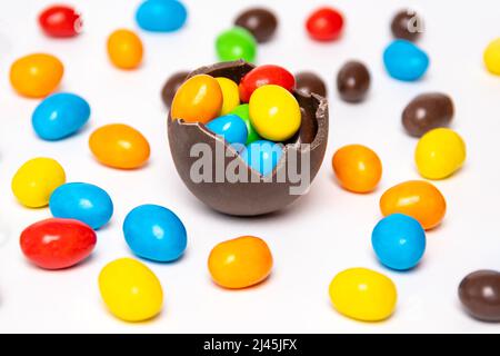 Half of a cracked chocolate egg filled with multi-colored candies on a white background with multi-colored candies, top view. Treats for children. Swe Stock Photo
