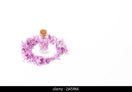 Glass bottle standing on a white background surrounded by lilac flower petals in the shape of a circle. Idea for floral fragrances. Stock Photo