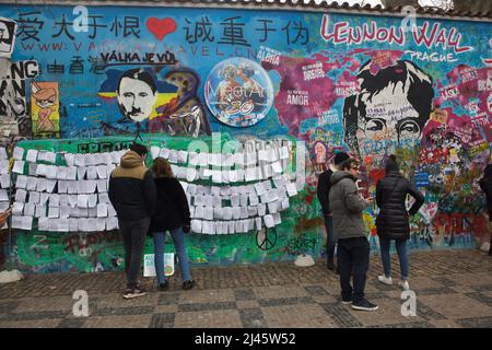 Russian president Vladimir Putin depicted next to John Lennon on the Lennon Wall (Lennonova zeď) in Prague, Czech Republic. President Vladimir Putin is depicted with characteristic haircut and toothbrush mustache of Adolf Hitler placed on the blooded map of Ukraine to protest against the Russian invasion of Ukraine in 2022. The Czech inscription over the Putin's head 'Válka je vůl' (Literary means: War is an ox) is a line from a popular song of Czech rock band 'Synkopy 61'. Tourists pictured on 2 April 2022 read antiwar lyrics placed next to the Lennon Wall. Stock Photo