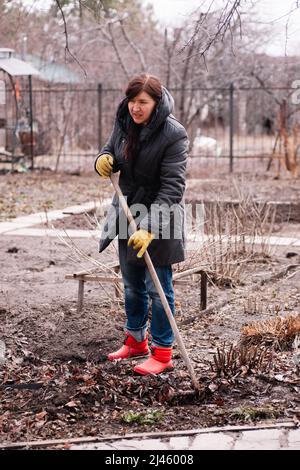 Tired woman farmworker gathering old fallen autumn dry leaves, foliage and cleaning area using rake in backyard. Close Stock Photo