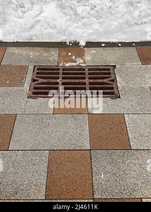 Rusty storm drain grate on tiled road and snowdrift that will melt in spring Stock Photo