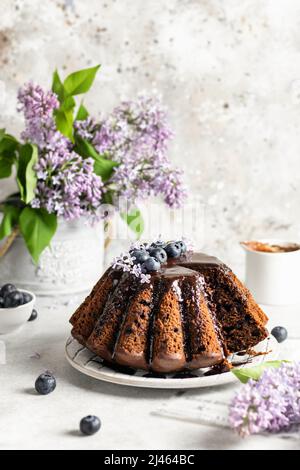 Sliced Chocolate cake decorated with chocolate icing, blueberries and lilac flowers on top on white textured background, spring composition with lilac Stock Photo