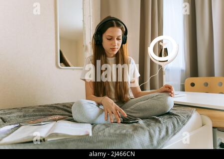 Young teenage girl in headphones and with smartphone is doing homework sitting on bed. A girl of generation Z with long hair, she is studying intently Stock Photo