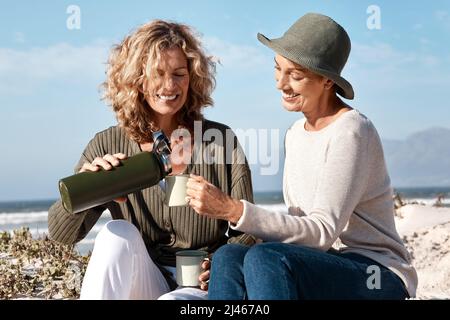 Can I top you up. Cropped shot of two attractive mature women enjoying some coffee while sitting on the beach. Stock Photo
