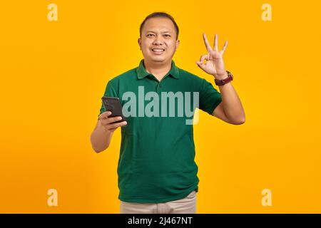 Smiling young man using smartphone and doing okay gesture on yellow background Stock Photo
