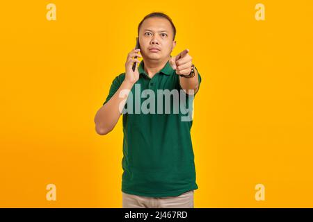 Angry Asian young man talking on smartphone and doing warning sign isolated over yellow background Stock Photo