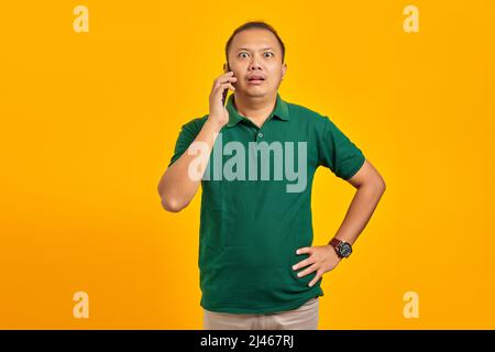 Surprised young Asian man talking on mobile and putting hands on waist on yellow background Stock Photo