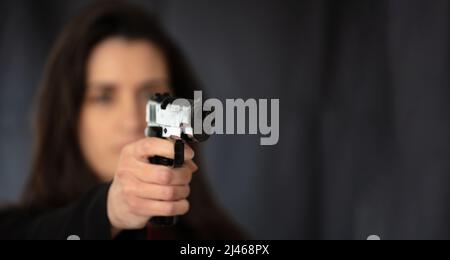 Woman holding a gun in hand pointing at camera. Female secret police agent, killer or spy long hair in dark background, copy space. Selective focus Stock Photo