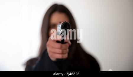 Woman holding a gun in hand pointing at camera. Female secret police agent, killer or spy dark long hair, copy space. Selective focus Stock Photo