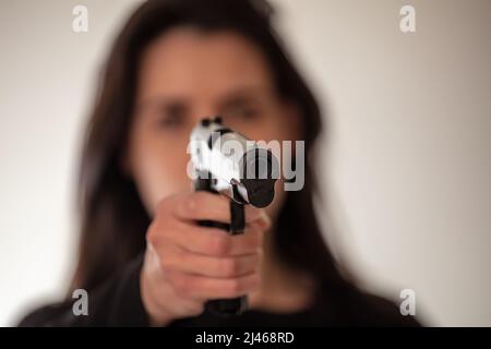 Woman holding a gun in hand pointing at camera. Brunette female secret police agent, killer or spy dark long hair. Selective focus Stock Photo