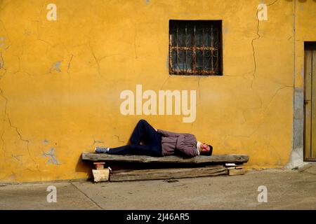 A man sleeps happily on a rough wooden bench in the sunshine beside a yellow wall in Becerril de Campos, near Palancia, Castile and León, Spain Stock Photo