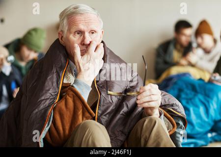 Portrait of Caucasian distressed senior man hiding in refugee shelter and staring in space vacantly Stock Photo