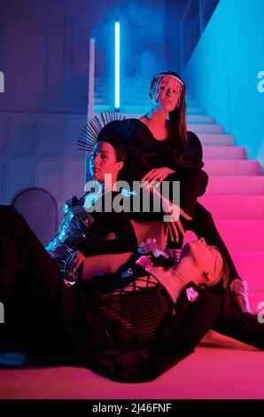 Group of three youthful vogue dance performers in posh attire posing on staircase of modern loft studio lit with pink and blue neon lights Stock Photo