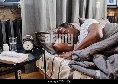 Tired African American male pensioner napping under gray blanket in bed in front of small table with alarm clock and medicaments Stock Photo