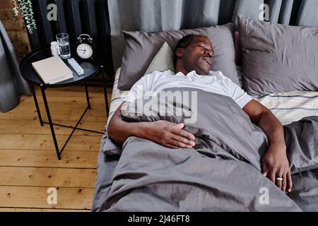 Tired senior man of African ethnicity sleeping under grey blanket with his head on pillow while having rest on comfortable double bed Stock Photo