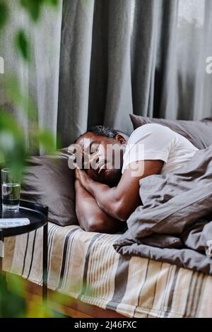Tired mature African American man sleeping peacefully in bed in the morning in front of small black table with glass of water Stock Photo
