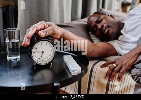 Hand of senior African American sleepy man pressing buzz bell of alarm clock on small round table in the morning while staying in bed Stock Photo