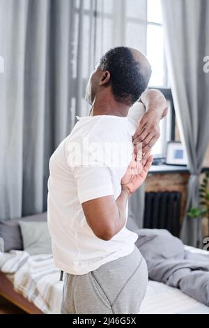 African American elderly man in pajamas doing stretching exercise for spinal cord while standing against bed in front of camera Stock Photo