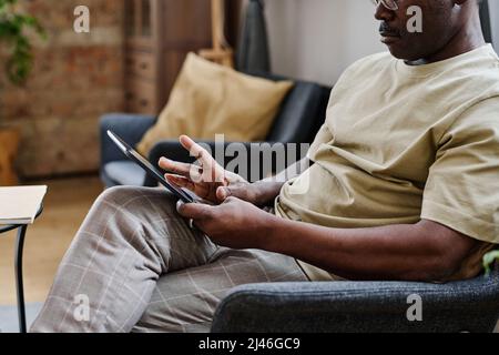 Close-up of contemporary senior man in pajamas using digital tablet while sitting in armchair at leisure in living room of his apartment Stock Photo