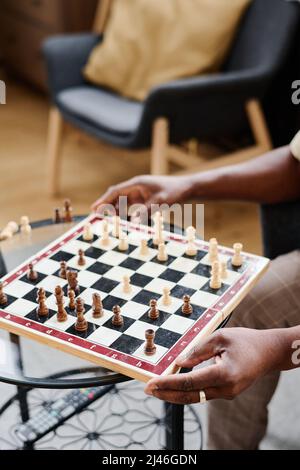 Hands of senior African American man holding chess board with black and white wooden figures while sitting by small glass table Stock Photo