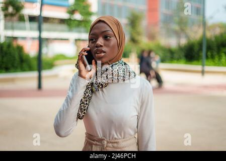 Optimistic African American female in stylish outfit and hijab having conversation on cellphone in urban park Stock Photo