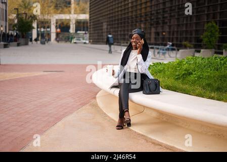 Joyful African American lady in trendy smart casual wear and Muslim headscarf sitting with cup of takeaway drink in urban park and talking on mobile phone Stock Photo