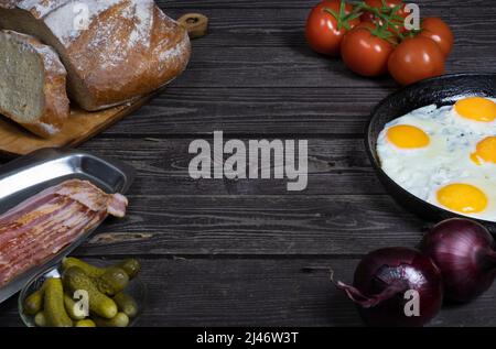 rustic fried eggs on a dark wooden table with tomatoes,, onion, beacon, bread and pickled gherkins. in dark key with copy space Stock Photo