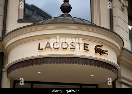 Lacoste lettering and logo on the facade of a building. Illuminated sign as advertisement on a retail store. Luxury fashion brand  with a crocodile. Stock Photo