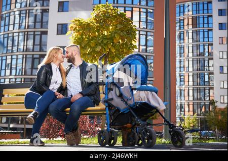 Happy family sitting with unrecognizable baby in blue pram, hugging, enjoying together. Side view of young parents sitting on bench with stroller. Modern residential buildings on background. Stock Photo