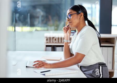 Hello, how can I help you. Shot of an attractive young businesswoman sitting alone in her office and wearing a headset while using her computer. Stock Photo