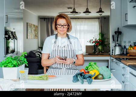 Woman using phone and looking for recipe online for cooking dinner in kitchen at home. Watching tutorial video while preparing healthy dish with fish Stock Photo