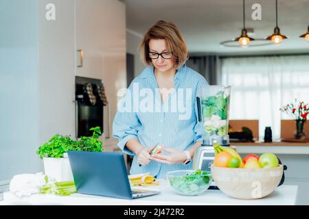 Middle age woman watching a cooking tutorial video course on laptop while preparing a meal in a kitchen. Woman making green smoothie. Online recipe. H Stock Photo