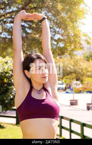 A young caucasian female athlete doing back stretching exercises by raising her arms with joined hands. The background is out of focus and the framing Stock Photo