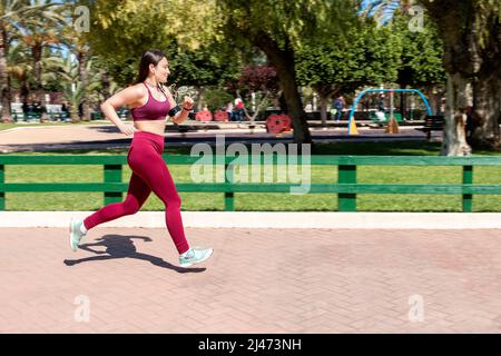 Young Caucasian female athlete dressed in sportswear running through a park with a sweeping effect. The girl is wearing headphones, activity wristband Stock Photo