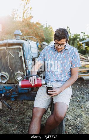 Young latin man serving mate with a red thermo, in a natural space, beside an old little blue tractor. Latin beverage. Stock Photo