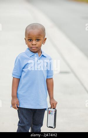 A little boy standing outside holding a cell phone and looking sad Stock Photo