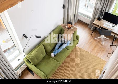 High angle view of the happy businessman working remotely in relaxed pose, man spends leisure time online in social. Optimistic man lying on the couch and using laptop computer for messaging Stock Photo