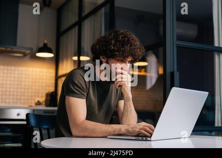 Side view of thoughtful young man programming on laptop sitting at table in kitchen room with modern interior. Stock Photo