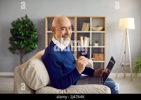 Happy, smiling senior man sitting on couch at home and using his laptop computer Stock Photo