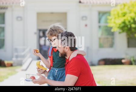 Father and son walking trough school park. Healthy school breakfast for child. Food for lunch, lunchboxes with fruits. Outdoor school. Stock Photo