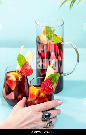 Female hand holding a glass with traditional Spanish Sangria cocktail decorated with pieces of fresh strawberry, pinch of mint and eco-paper straw. Stock Photo